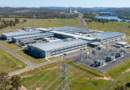 Aerial view of a large data center building with adjacent electrical substation and power transmission lines in a rural area.