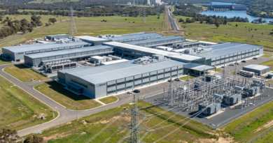 Aerial view of a large data center building with adjacent electrical substation and power transmission lines in a rural area.