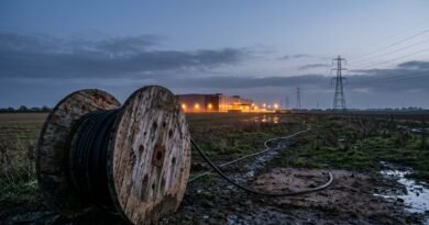 Large wooden cable drum in a muddy Essex field with a black power cable that stops short of a distant lit data centre, beside electricity pylons at dusk