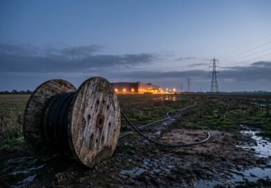 Large wooden cable drum in a muddy Essex field with a black power cable that stops short of a distant lit data centre, beside electricity pylons at dusk